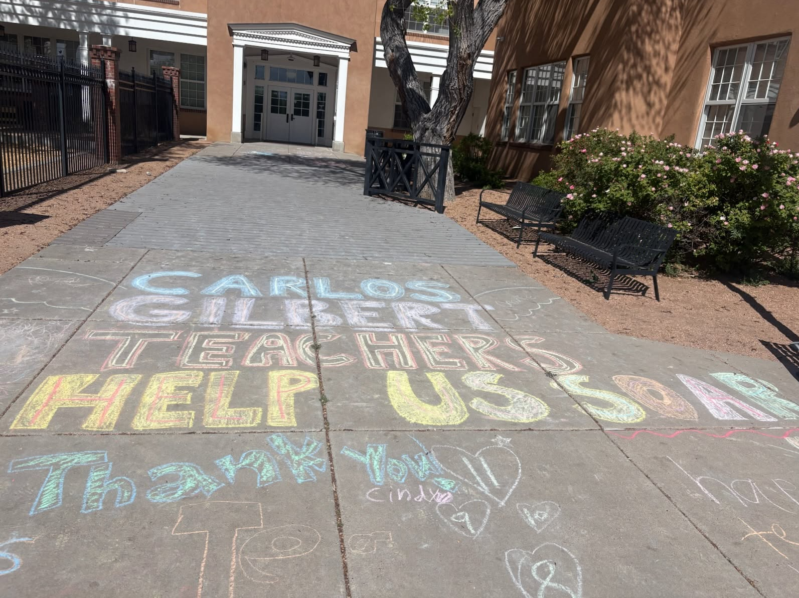 Sidewalk with sidewalk chalk reading "Carlos Gilbert Teacher Help Us" and other messages. Two benches, building, and trees in the background.