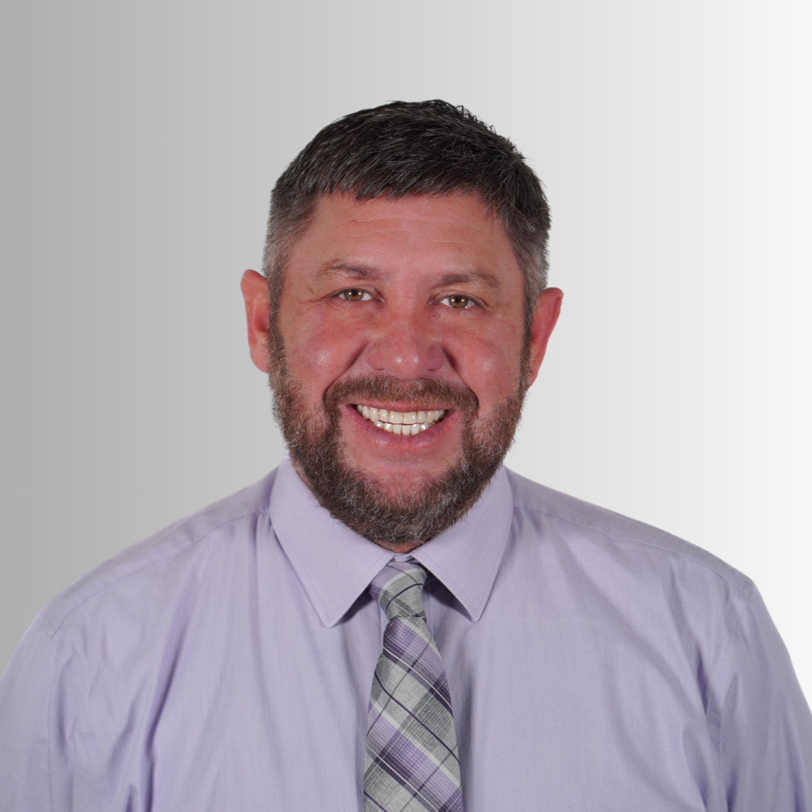A man in a purple shirt with a gray tie, short dark hair, and a beard smiles at the camera.