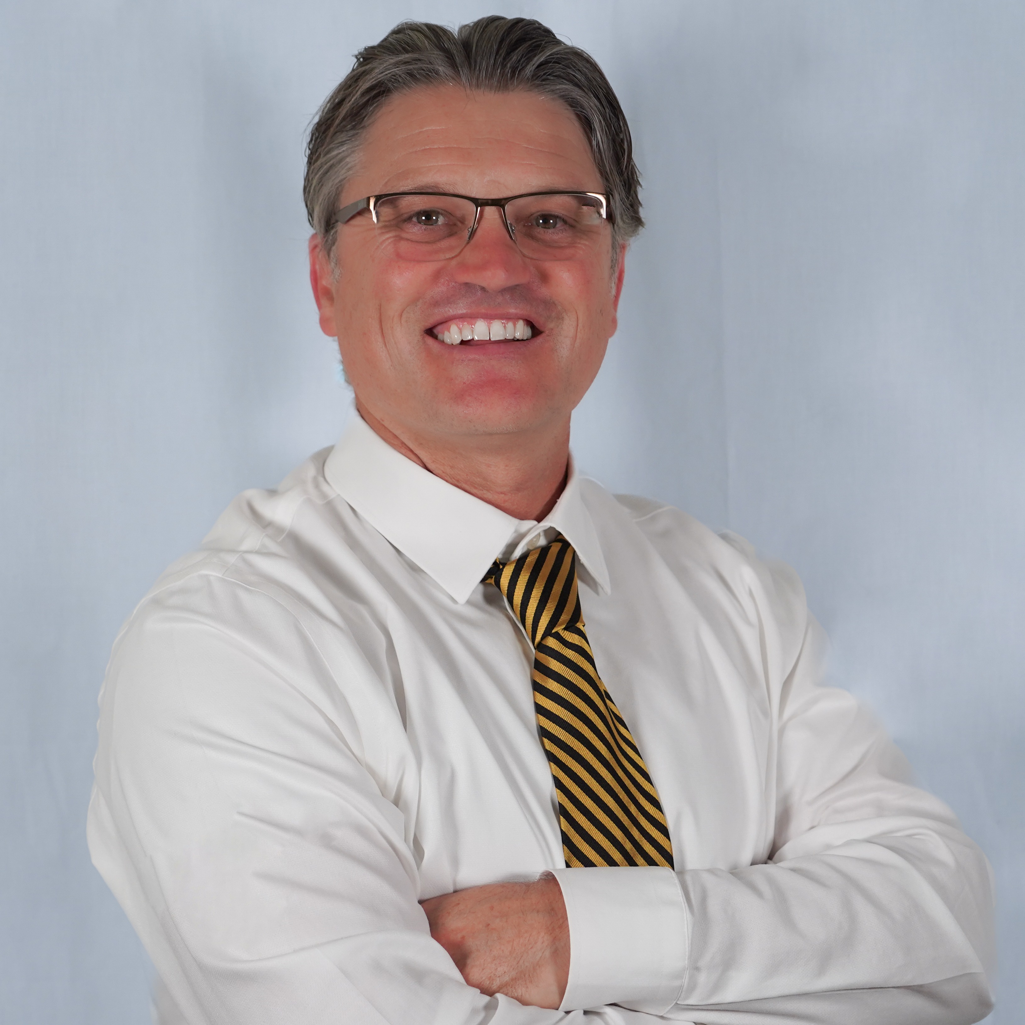 A man with glasses, a white shirt, and a black and yellow tie. He has his arms folded.