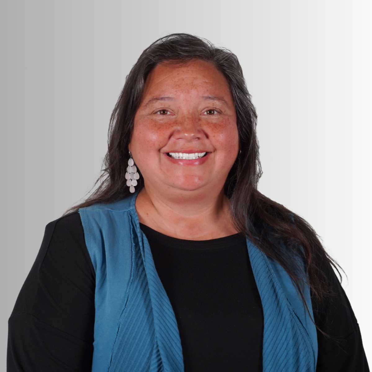 A woman with long hair and a blue vest smiles at the camera. She wears a black top and earrings.