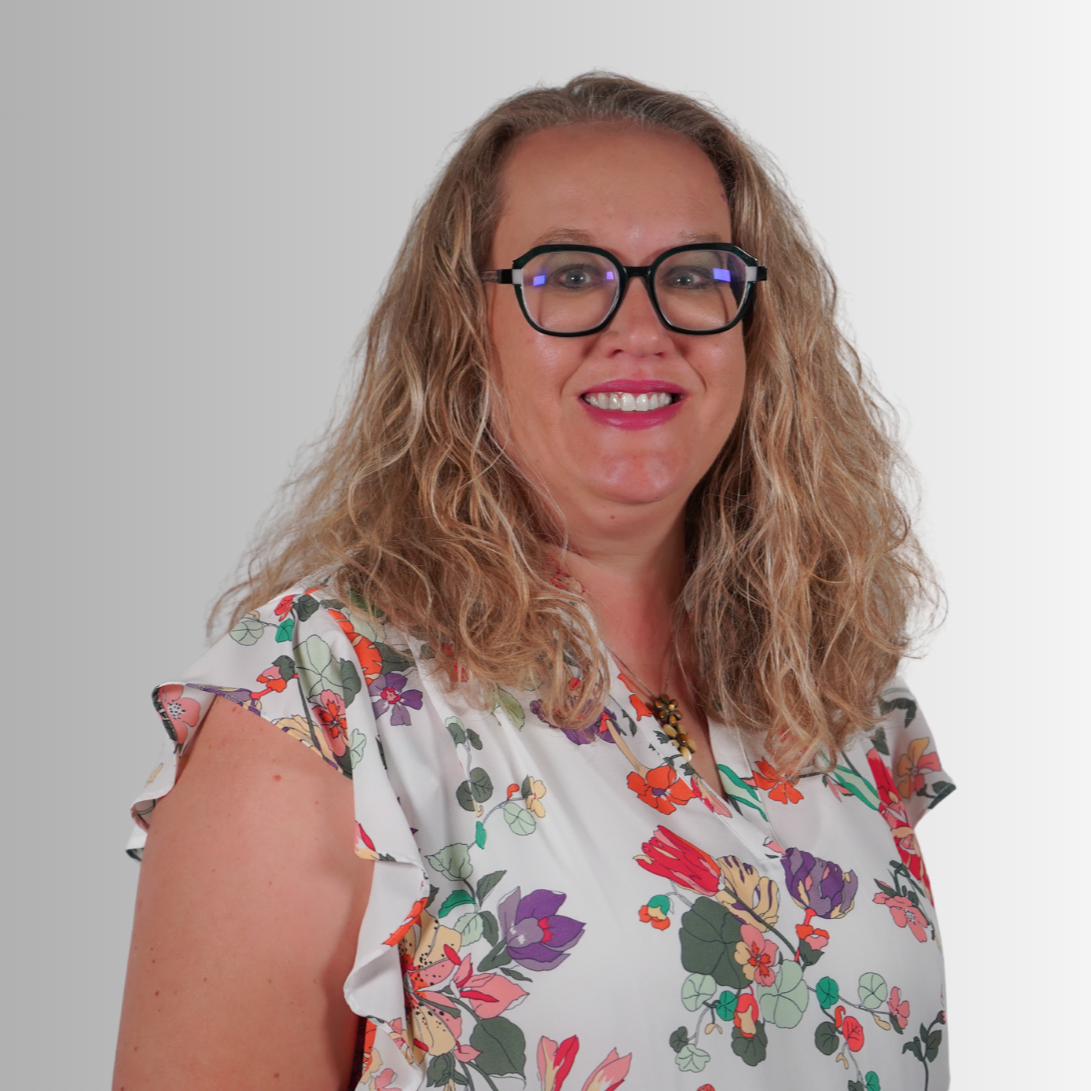 An adult female with long blonde hair smiles in front of a plain gray background. She wears a short-sleeved floral blouse and glasses.