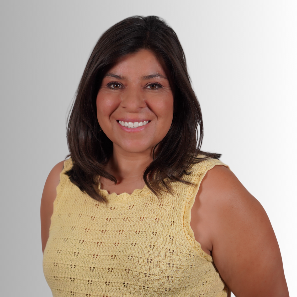A woman with dark hair smiles at the camera, wearing a yellow sleeveless top against a plain gray background.