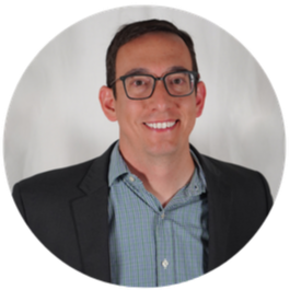 A man with glasses, wearing a blue checkered shirt and dark blazer, smiling with a white background.