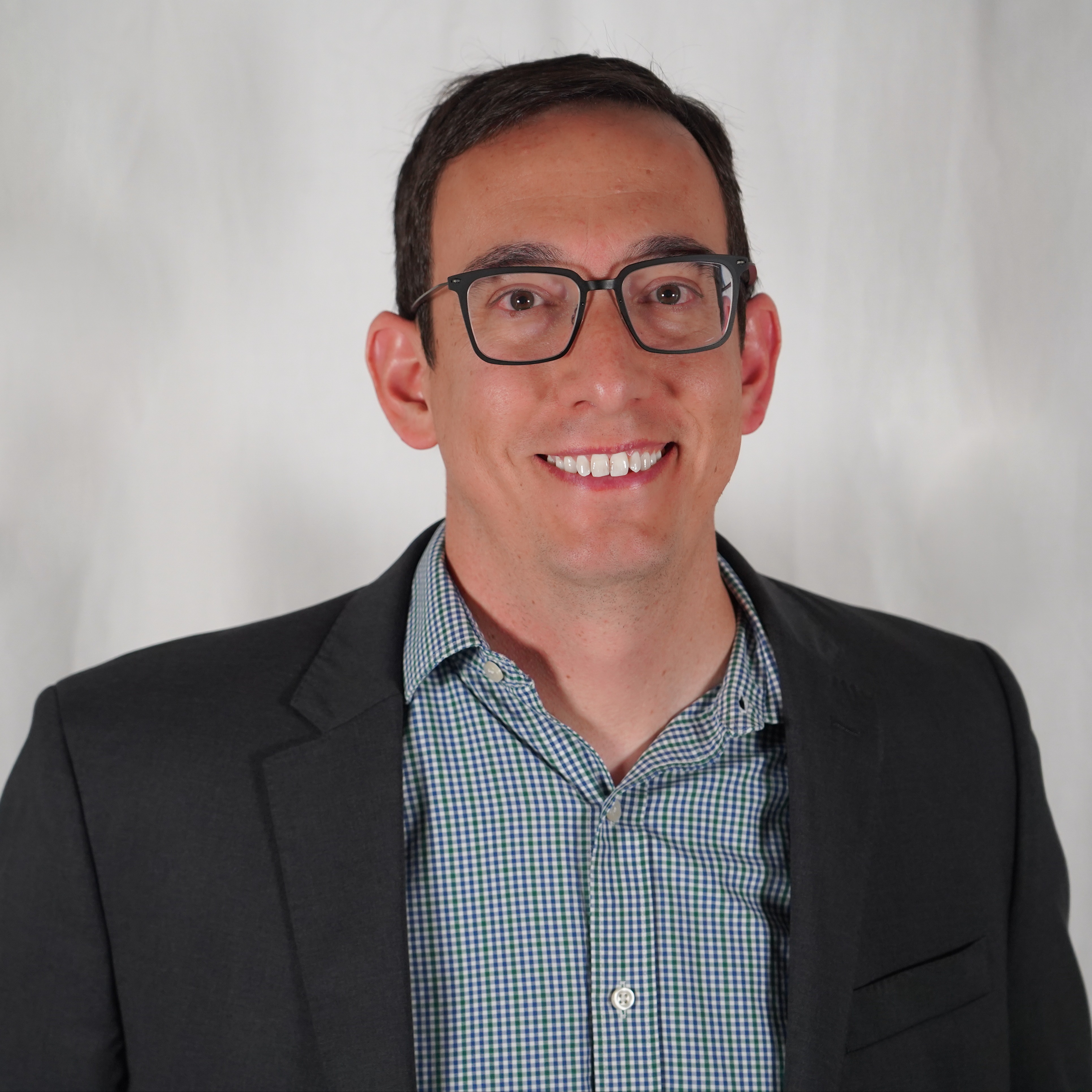 Man in glasses and a blue plaid shirt, wearing a black jacket, smiling. Background is a plain white curtain.