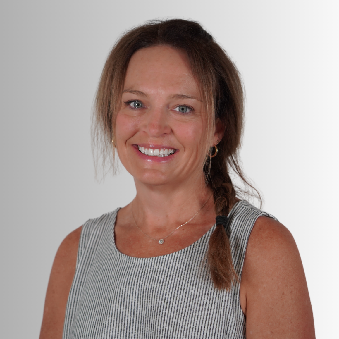 A woman with brown hair in a ponytail smiles for the camera, wearing a sleeveless gray top and a necklace.