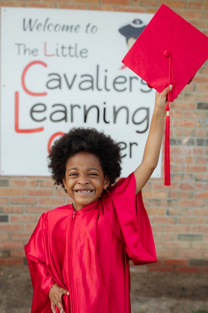A smiling young child wearing a bright red graduation gown stands outside the Little Cavaliers Learning Center. The child proudly raises a matching red graduation cap in the air while posing in front of a school sign. The image celebrates an early learning milestone and captures the excitement and joy of graduation day.