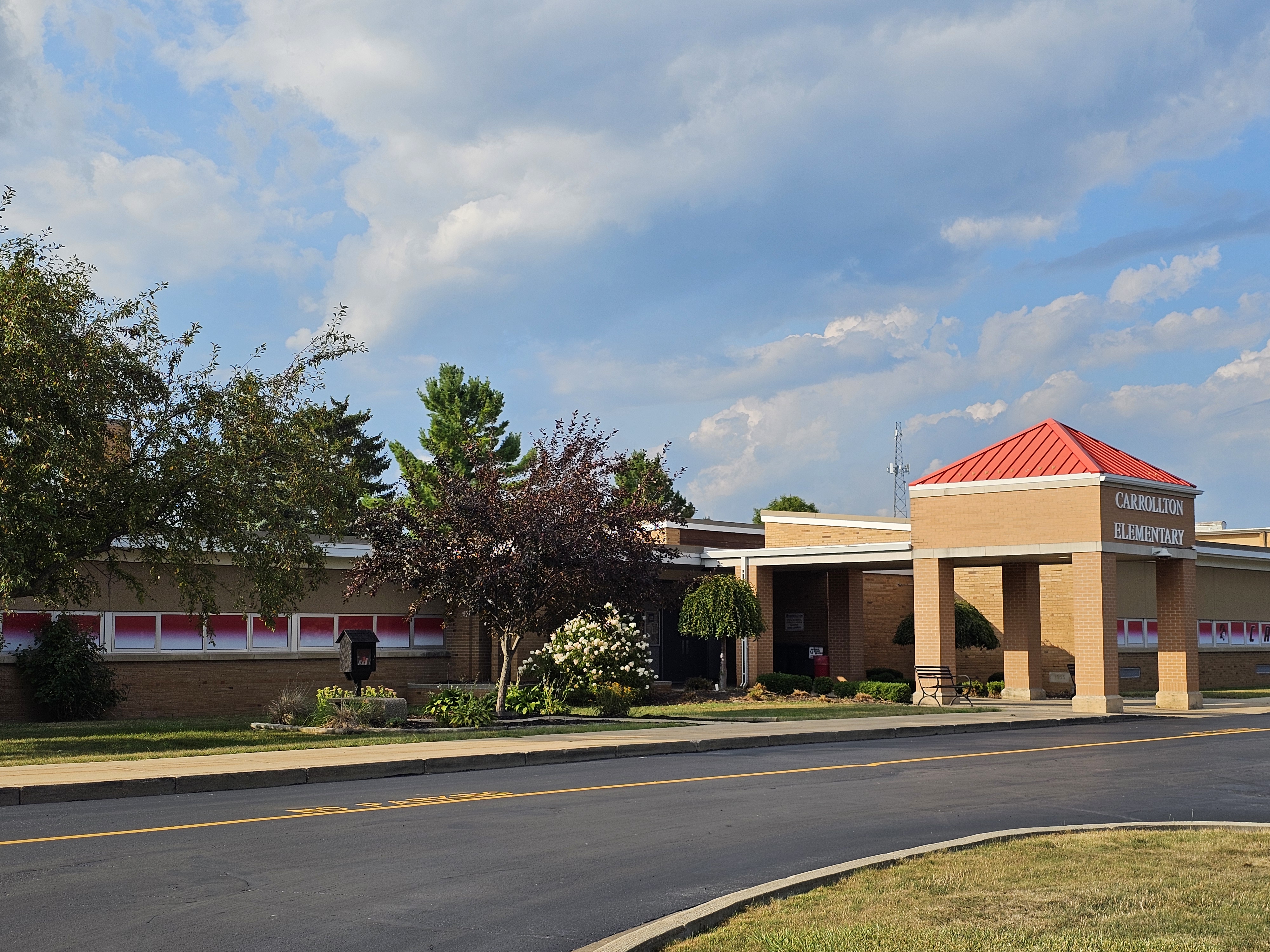 Exterior view of Carrollton Elementary School on a sunny day with large clouds in the sky. The brick school building features a covered entrance with a red roof and “Carrollton Elementary” signage. Trees, flowering bushes, landscaping, and the curved driveway are visible in front of the building.