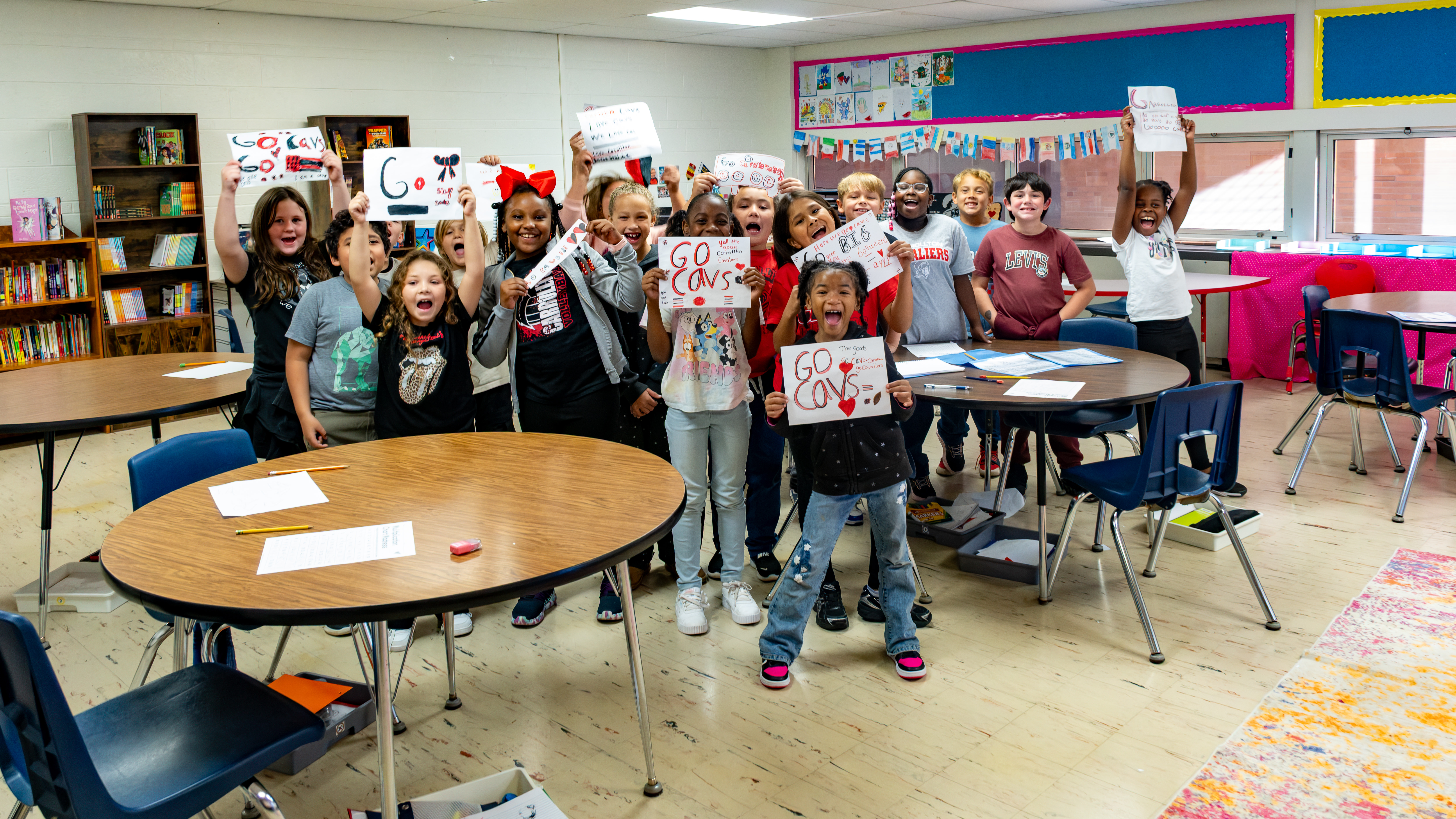 A group of enthusiastic Carrollton Elementary School students smile, cheer, and hold up handmade signs supporting the Cavaliers for Homecoming 2026. Several signs read “Go Cavs” and feature colorful drawings, hearts, and messages of school spirit. The students gather around classroom tables filled with art supplies, proudly showing off their creations and celebrating Cavalier pride.