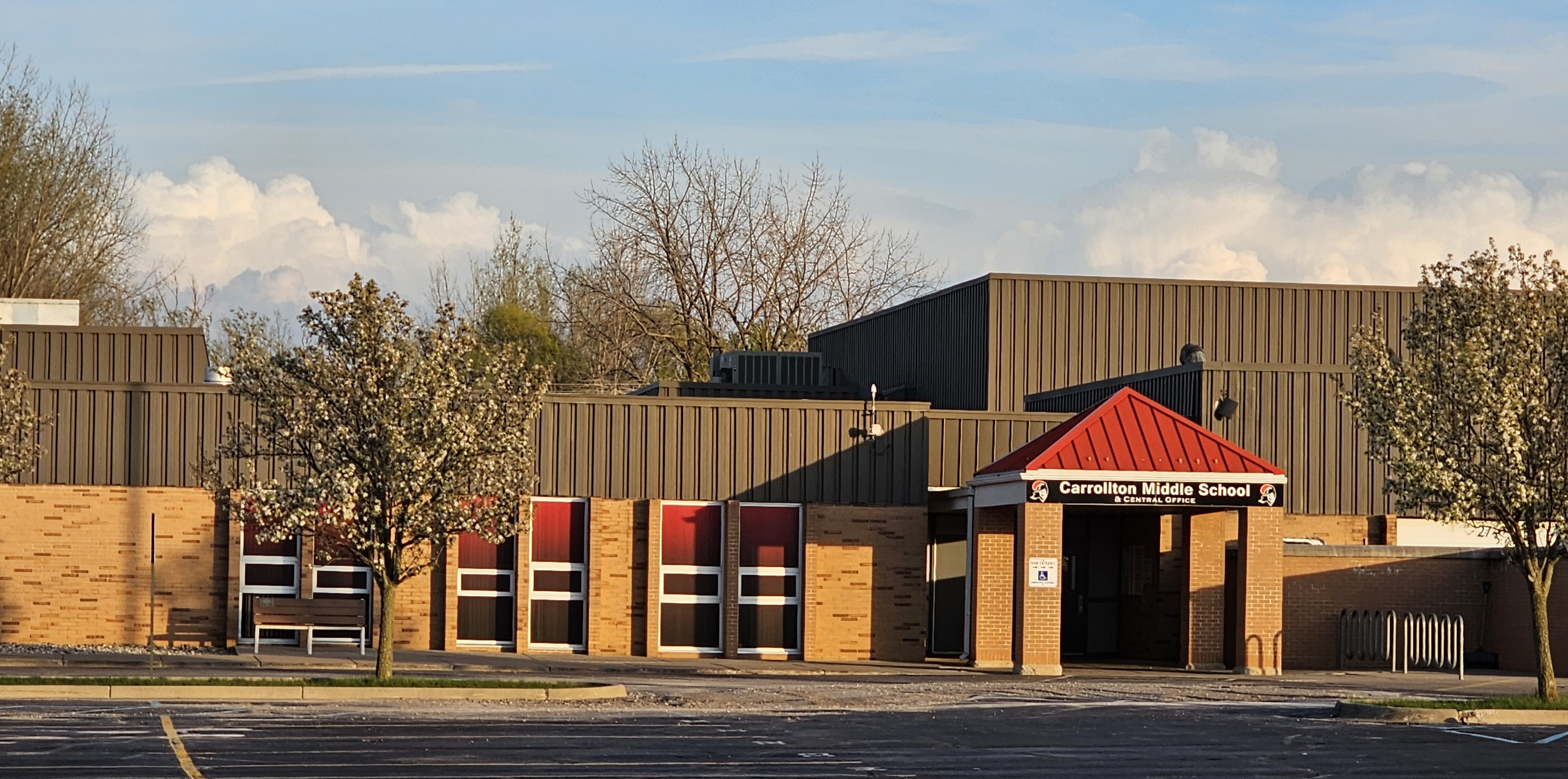 Front exterior of Carrollton Middle School and Central Office during springtime. The brick school building features a covered entrance with a red roof and signage reading “Carrollton Middle School &amp; Central Office.” Flowering trees frame the entrance, and the parking lot is visible in the foreground under a partly cloudy sky.