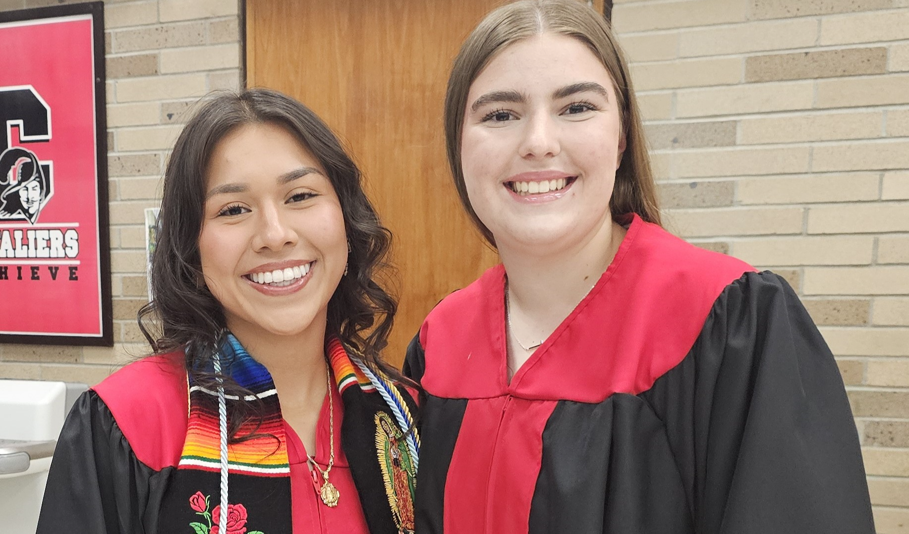 Two female students smile at camera while wearing red and black graduation gowns