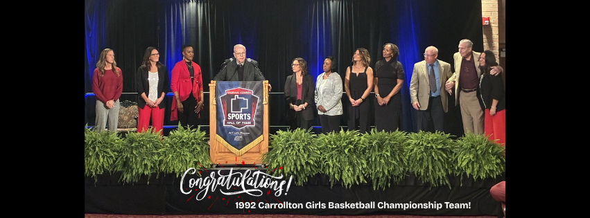 A group of adults stands on a stage behind a podium at an awards ceremony. The podium features a “Sports Hall of Fame” sign. One person is speaking into the microphone while the others stand in a line on either side, smiling and clapping. The stage is decorated with black curtains, blue lighting, and large green plants along the front. A banner at the bottom reads, “Congratulations! 1992 Carrollton Girls Basketball Championship Team!”