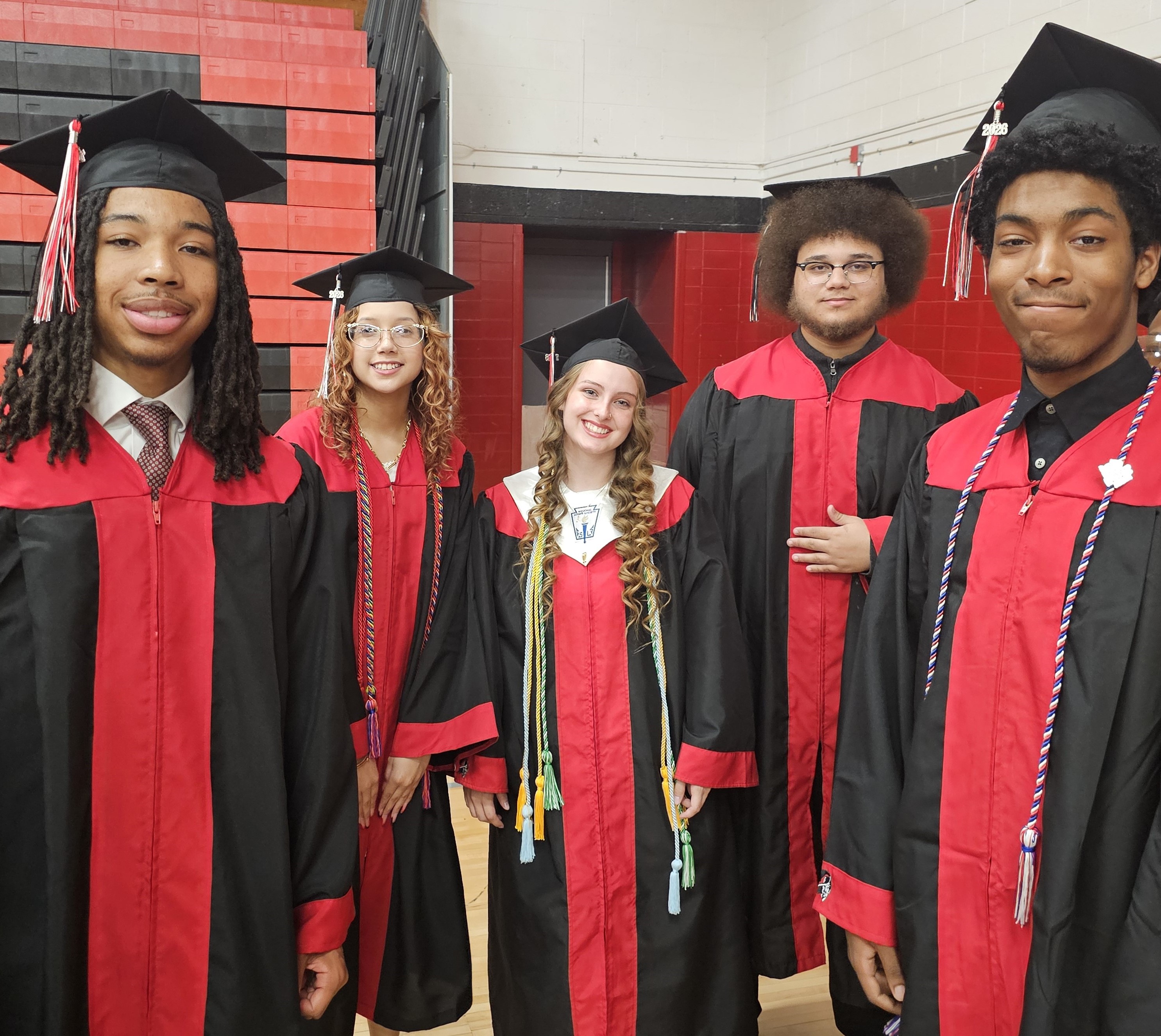 Group photo of graduating students wearing black caps and black graduation gowns with red panels, standing together inside a school gym decorated in Carrollton colors of red and black. Two students stand in the front smiling toward the camera, while additional graduates stand behind them, some wearing honor cords and graduation decorations. The students are gathered closely together, creating a celebratory Class of 2026 portrait highlighting pride and accomplishment.