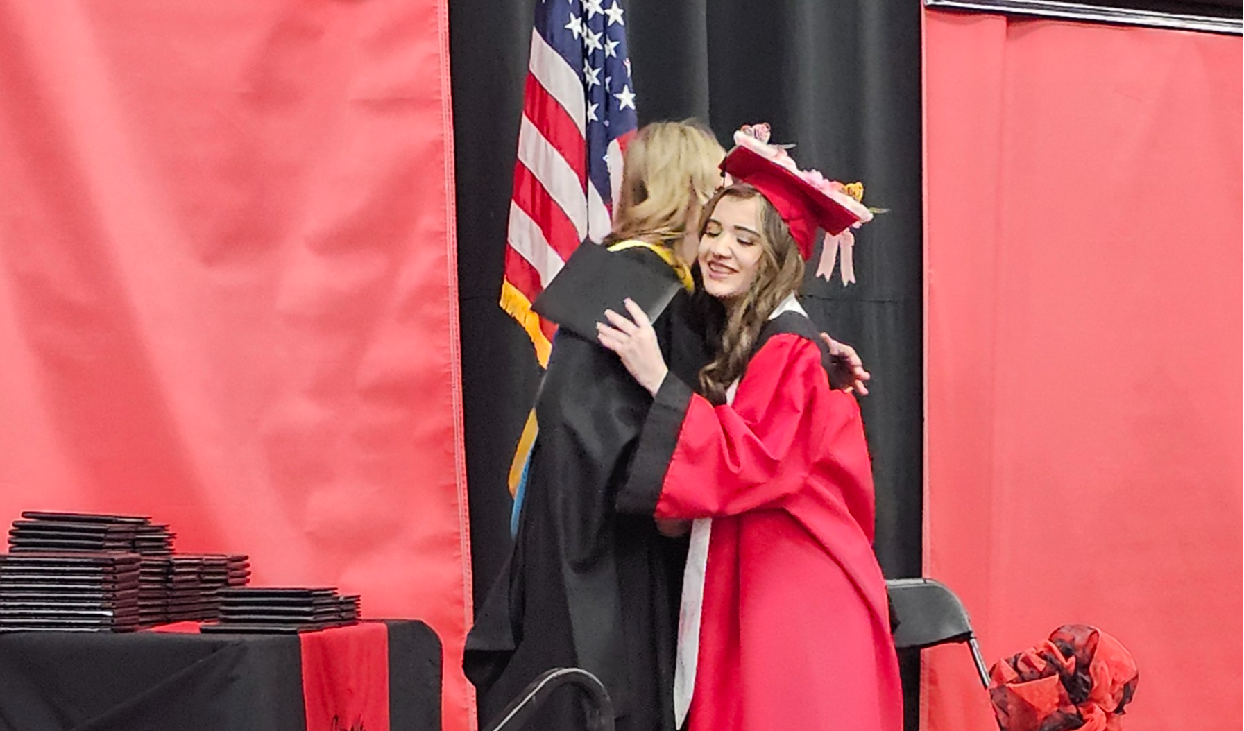A graduate in a red cap and gown hugs a staff member in a black robe on stage during a graduation ceremony. The stage is decorated with red and black drapes, an American flag, and ribbon bows. A stack of diploma covers is visible on a nearby table.