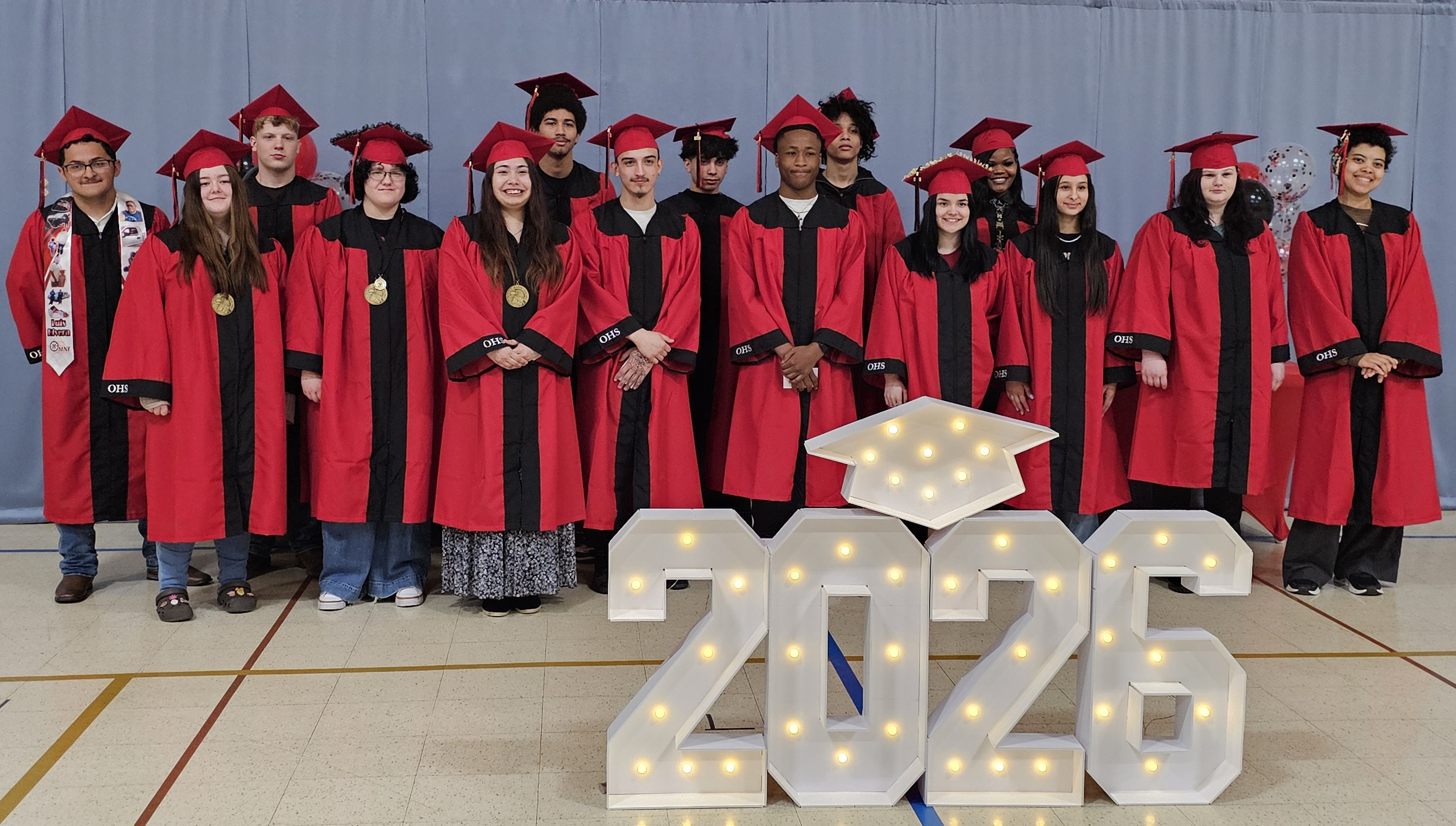 Group photo of the Omni High School Class of 2026 standing in two rows indoors in front of a gray backdrop. Students wear red graduation gowns and caps with black accents, with several graduates wearing medals or honor cords. A large illuminated “2026” display with a lighted graduation cap sits in front of the group. Balloons are visible in the background as graduates smile and pose for the commemorative photo.