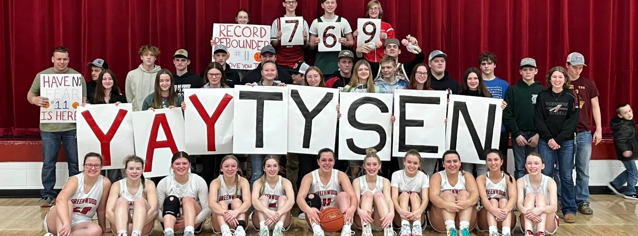 signs say yay tysen, students standing on stage with girls basketball team sitting on floor