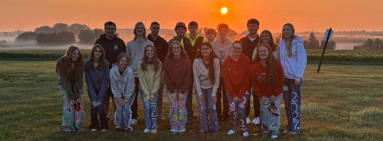 students standing in two rows in front of a sunrise for senior sunrise. fog, trees, grass, and sky