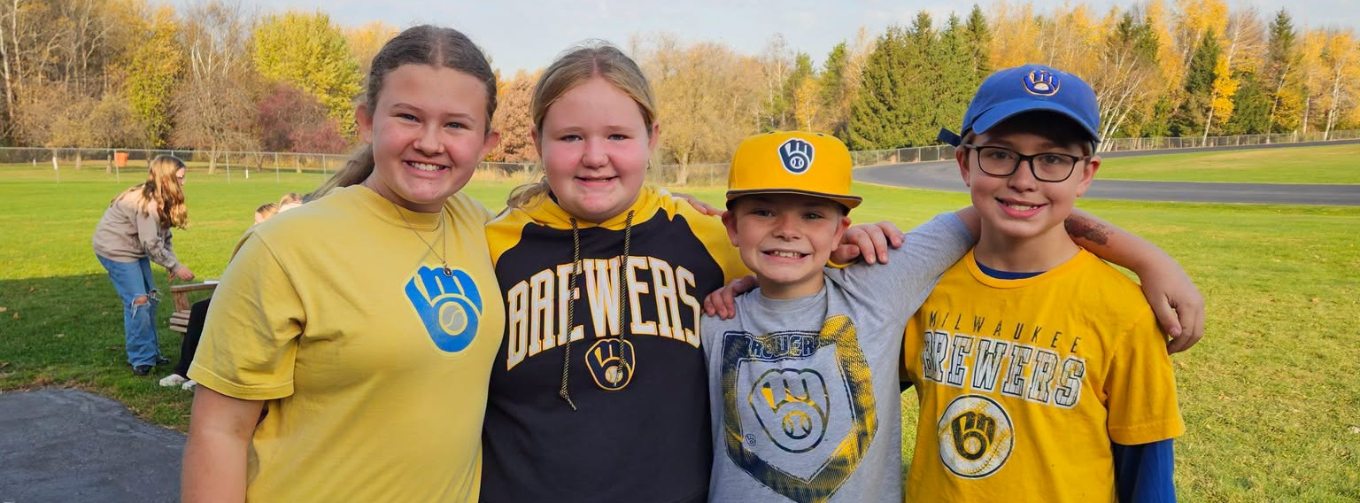 four students standing next to each other on the playground, wearing brewers apparel on a beautiful fall day