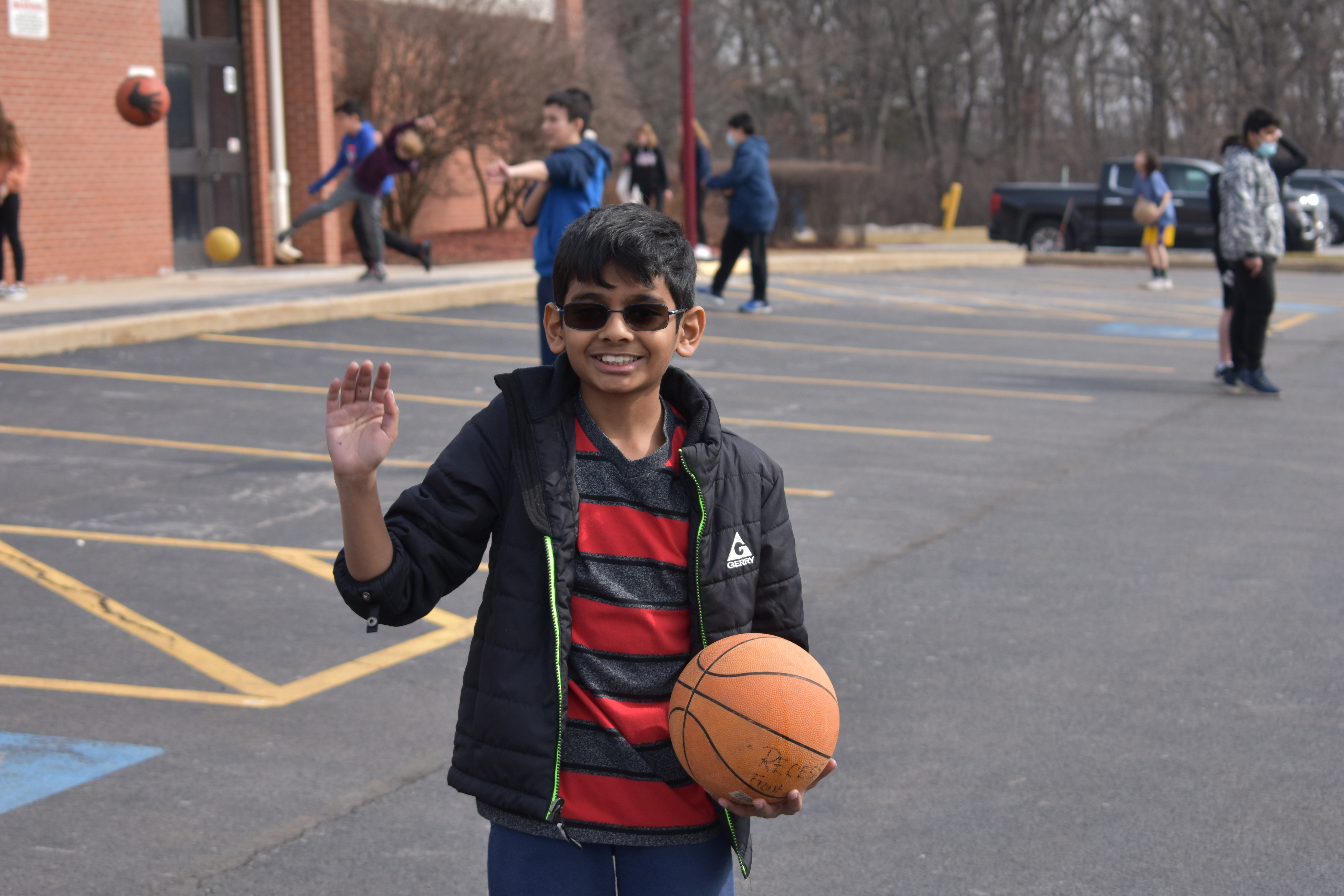 student holding a basketball and waving from the blacktop in front of the school