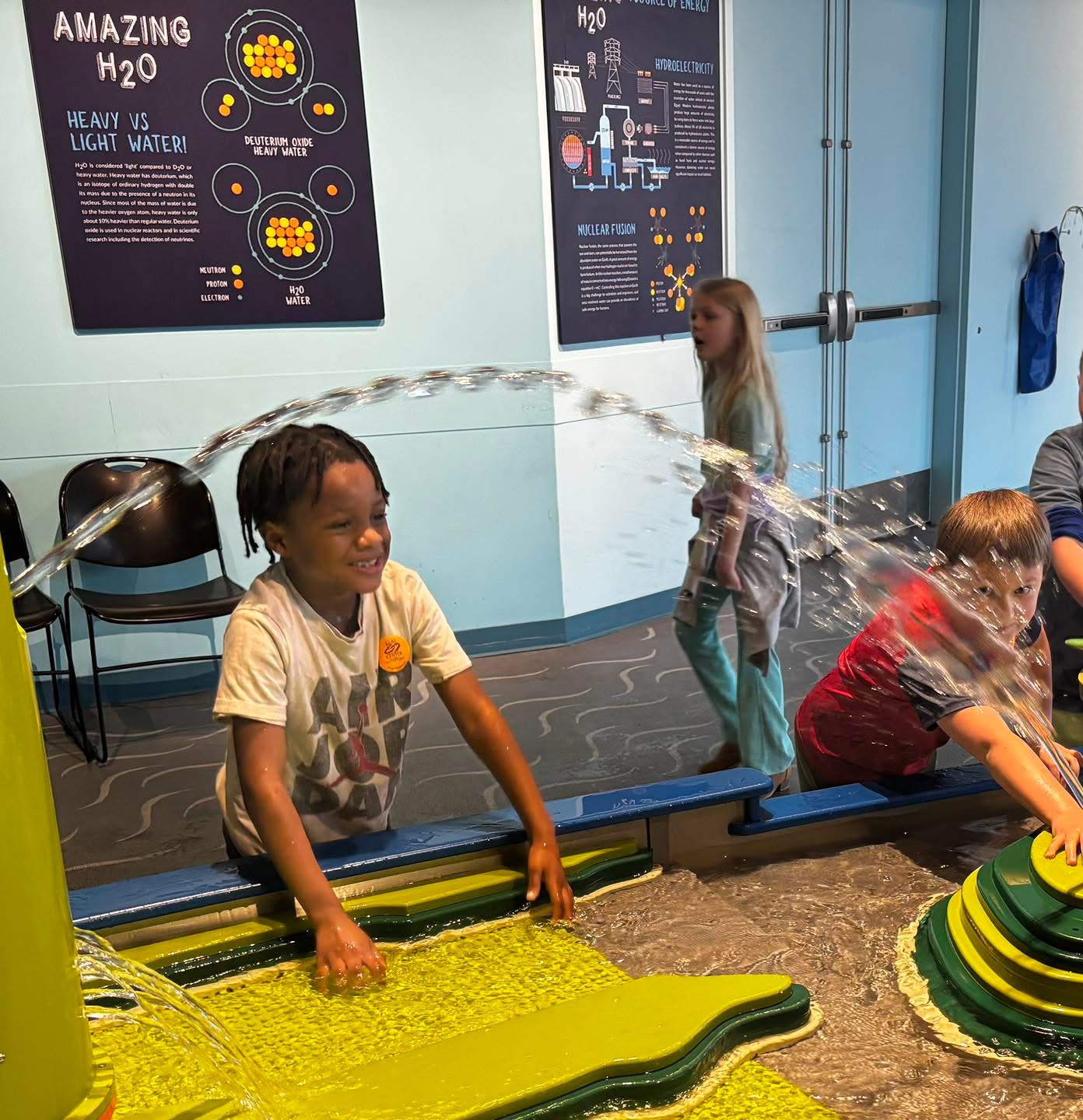 Children play with water fountains in an educational exhibit, with one child holding a water-themed poster.