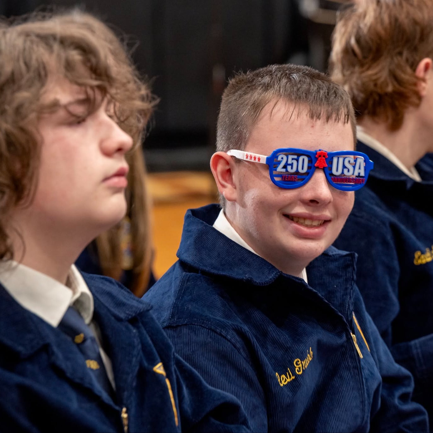 A group of students in navy blue jackets, some wearing glasses, sitting in a row. One wears USA-themed glasses.