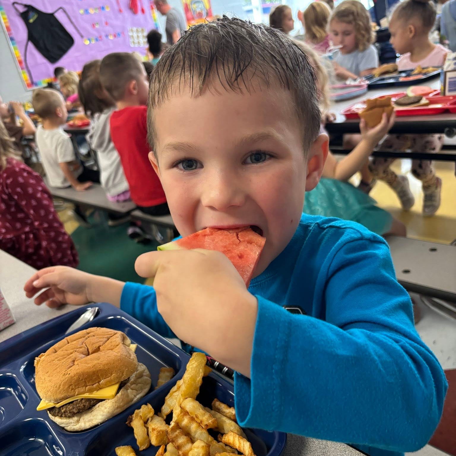 A child eating a hamburger, fries, and watermelon slice from a cafeteria tray, with other children in the background.
