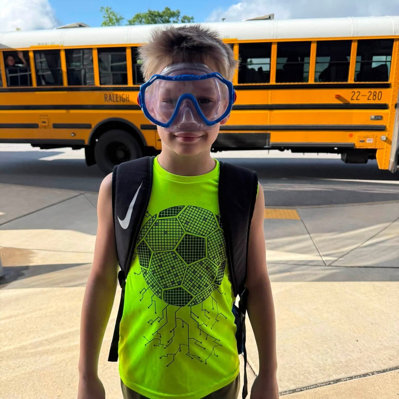 A child with goggles and a soccer ball-themed shirt stands in front of a yellow school bus.