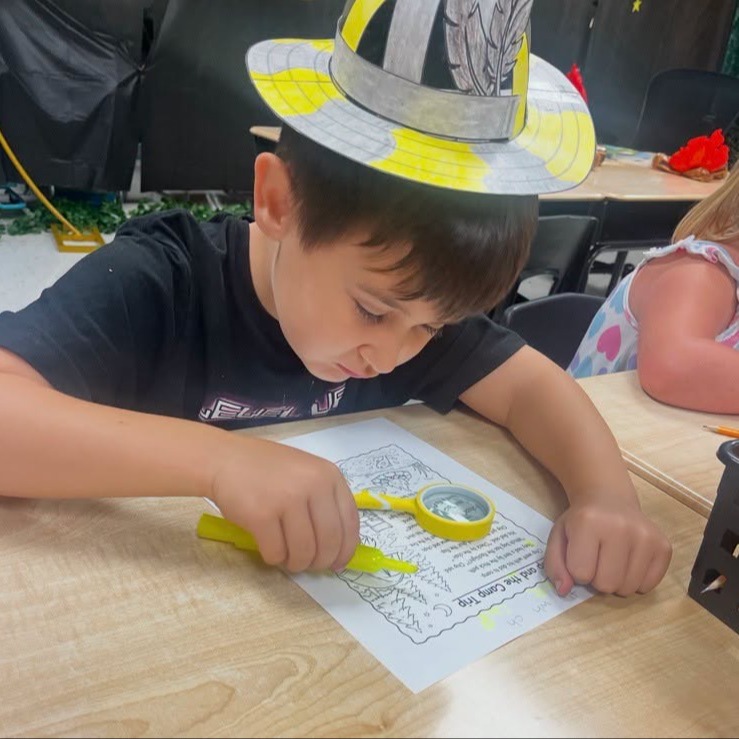 A child wearing a paper hat focuses on a desk, cutting paper with scissors. Behind, a girl sits at a desk.