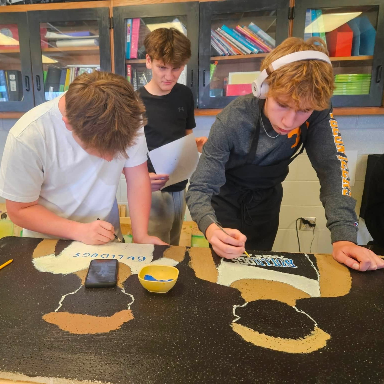 Three boys at a desk, one holding a paper, another with a pencil, painting a large black canvas.