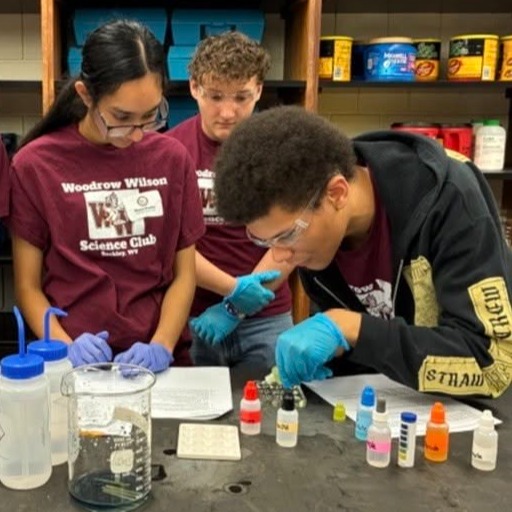 Four students wear lab coats and gloves and work at a lab table with various chemicals and beakers.