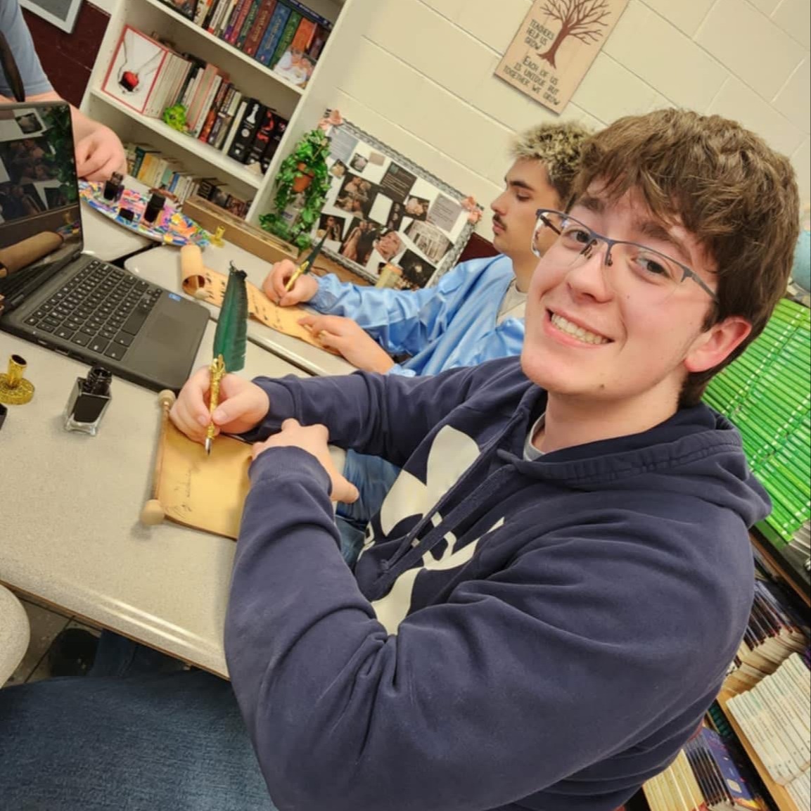 A person in a blue hoodie sits at a desk with a laptop and writes. Behind them, another person is seated, and a shelf with books is visible.