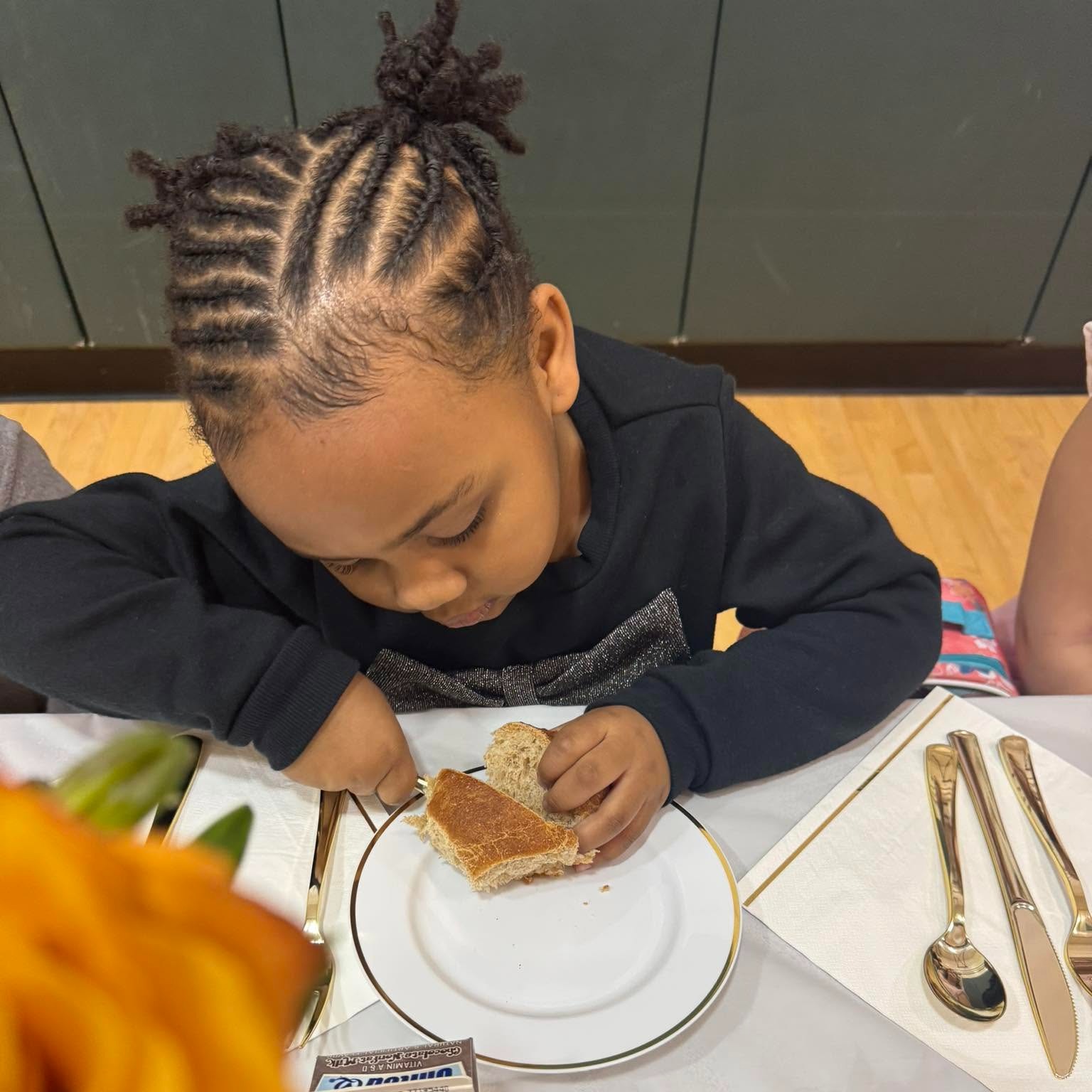A young child eats a sandwich at a table set with plates, utensils, and a rose.