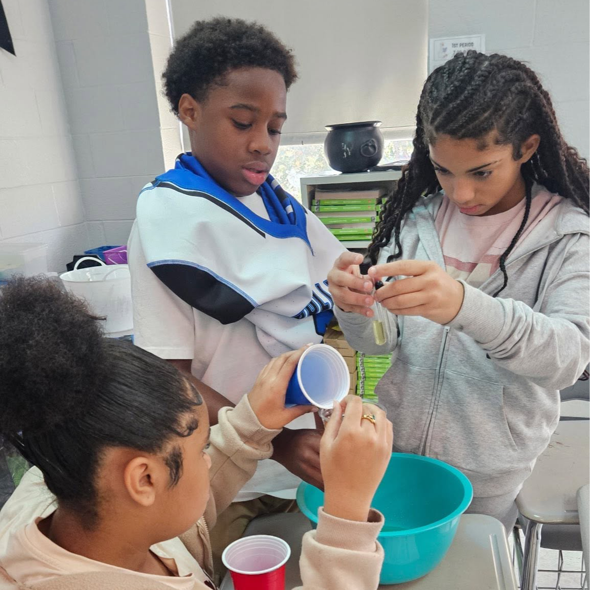 Three students in a classroom, two pouring liquid into a cup, one observing, with a blue bowl on the table and papers nearby.