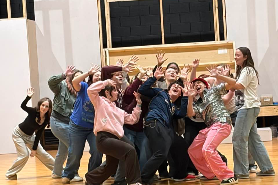A group of people on a wooden floor, smiling and dancing with hands raised, in front of a stage.