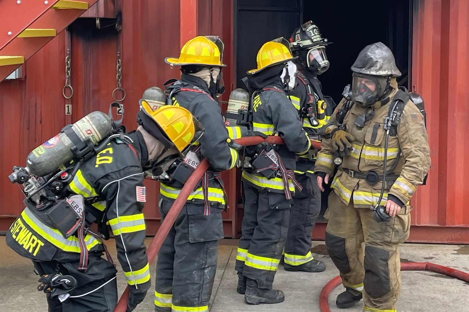 Firefighters in gear stand in front of a building. One holds a hose. Others wear helmets and masks. A staircase is on the left.