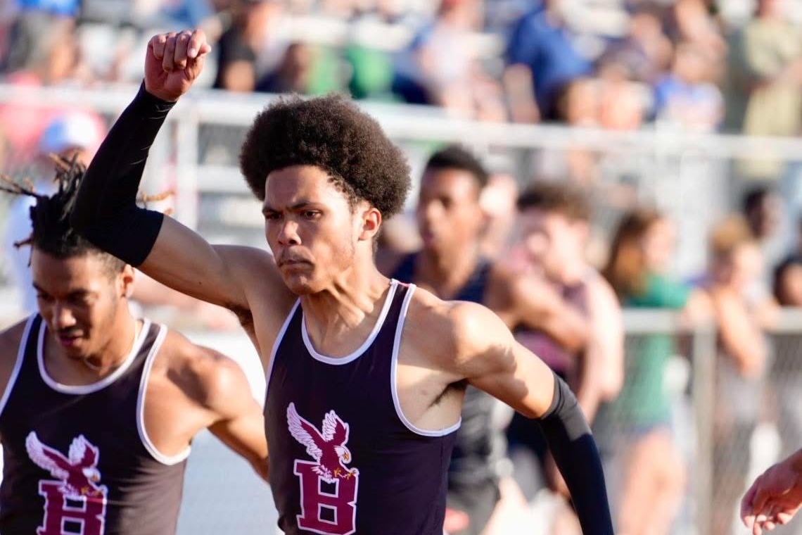 Two athletes run on a track, one with raised fist, both wearing black uniforms with pink "B" on chest.