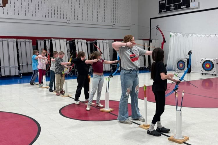 Several children practice archery in a gymnasium, with targets on the wall and a coach overseeing.