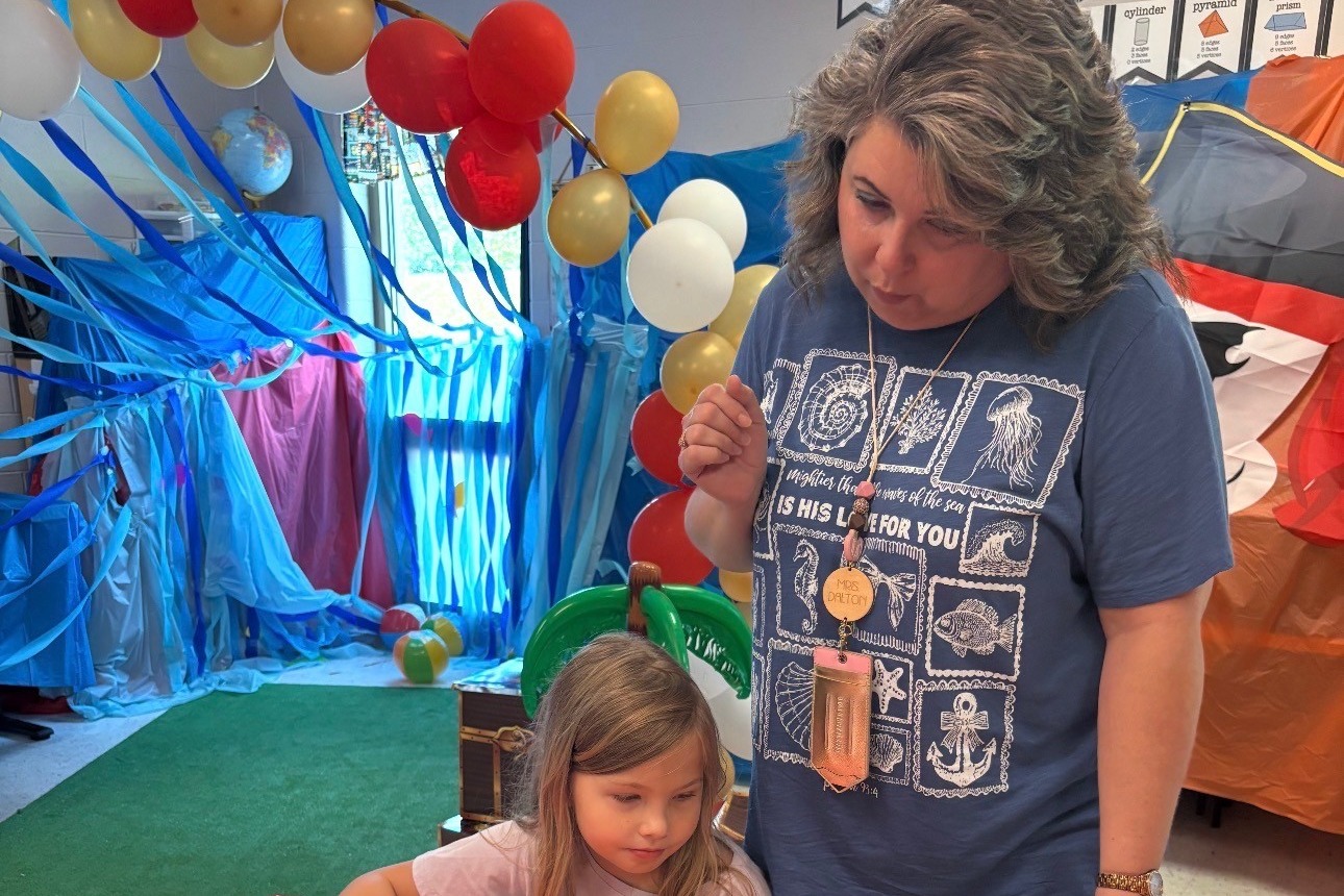 A woman helps a young girl with art supplies at a table with a red tablecloth.