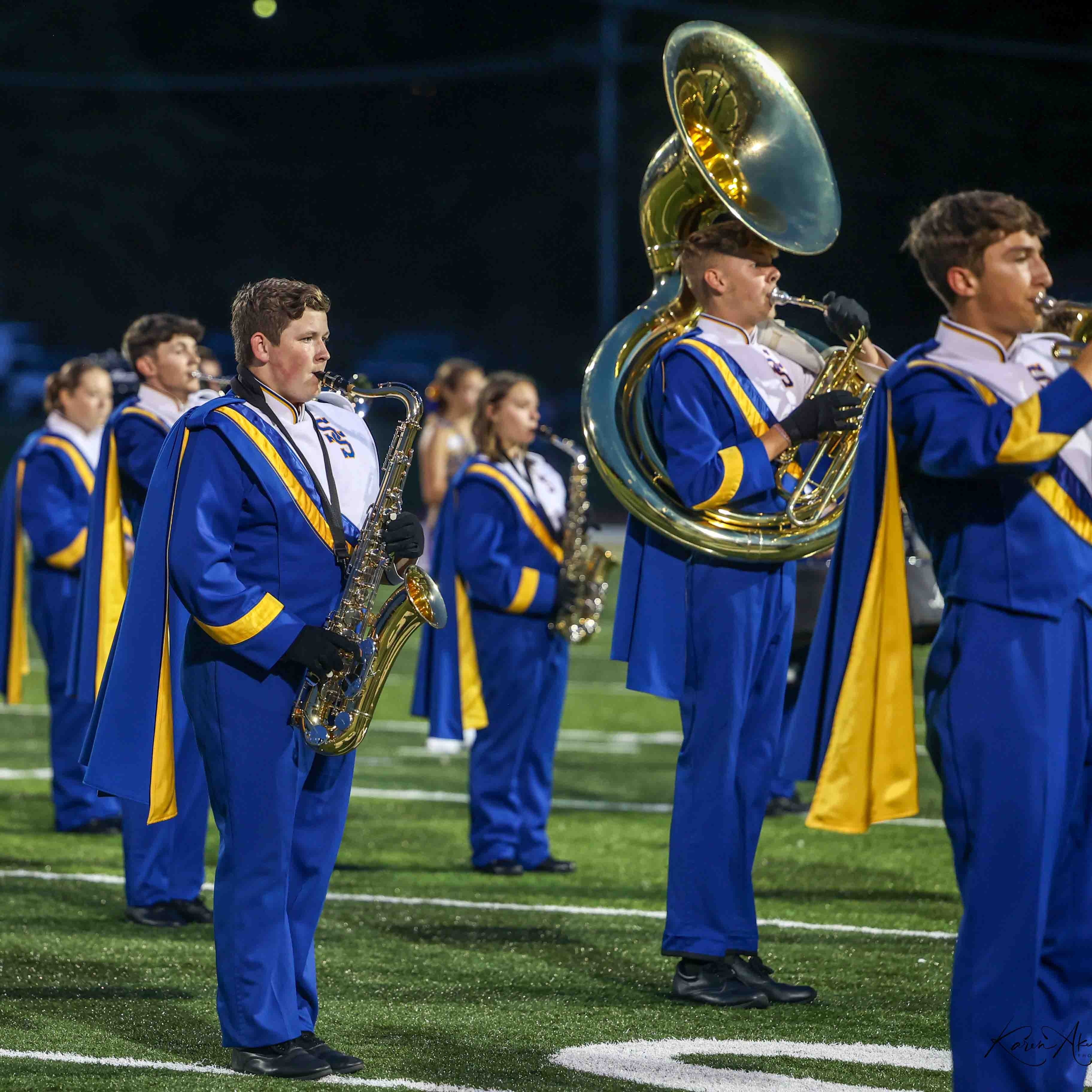 A group of musicians in blue and yellow uniforms play instruments on a football field.