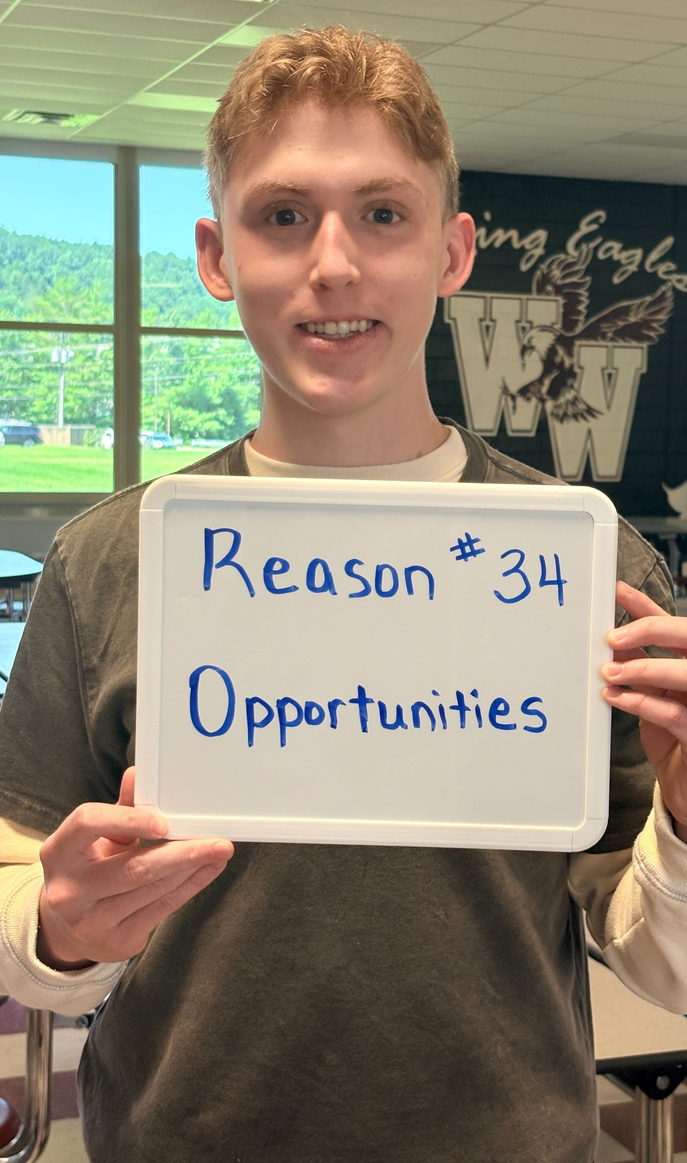 A smiling person holds a whiteboard that reads "Reason #34 Opportunities" in a school cafeteria with tables.