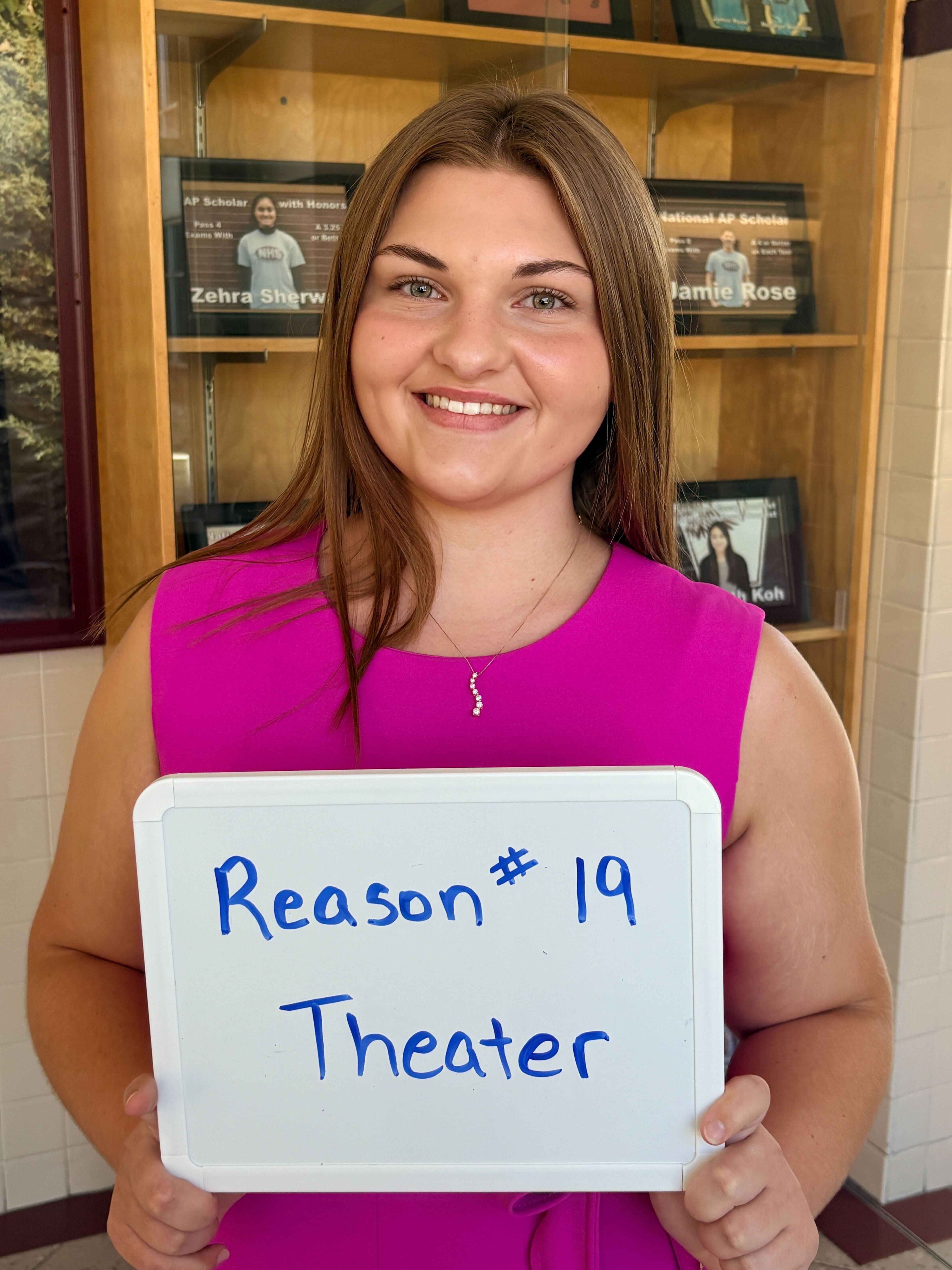 A smiling woman in a pink shirt holds a white sign that says "Reason #19 Theater" in front of a wooden bookshelf.