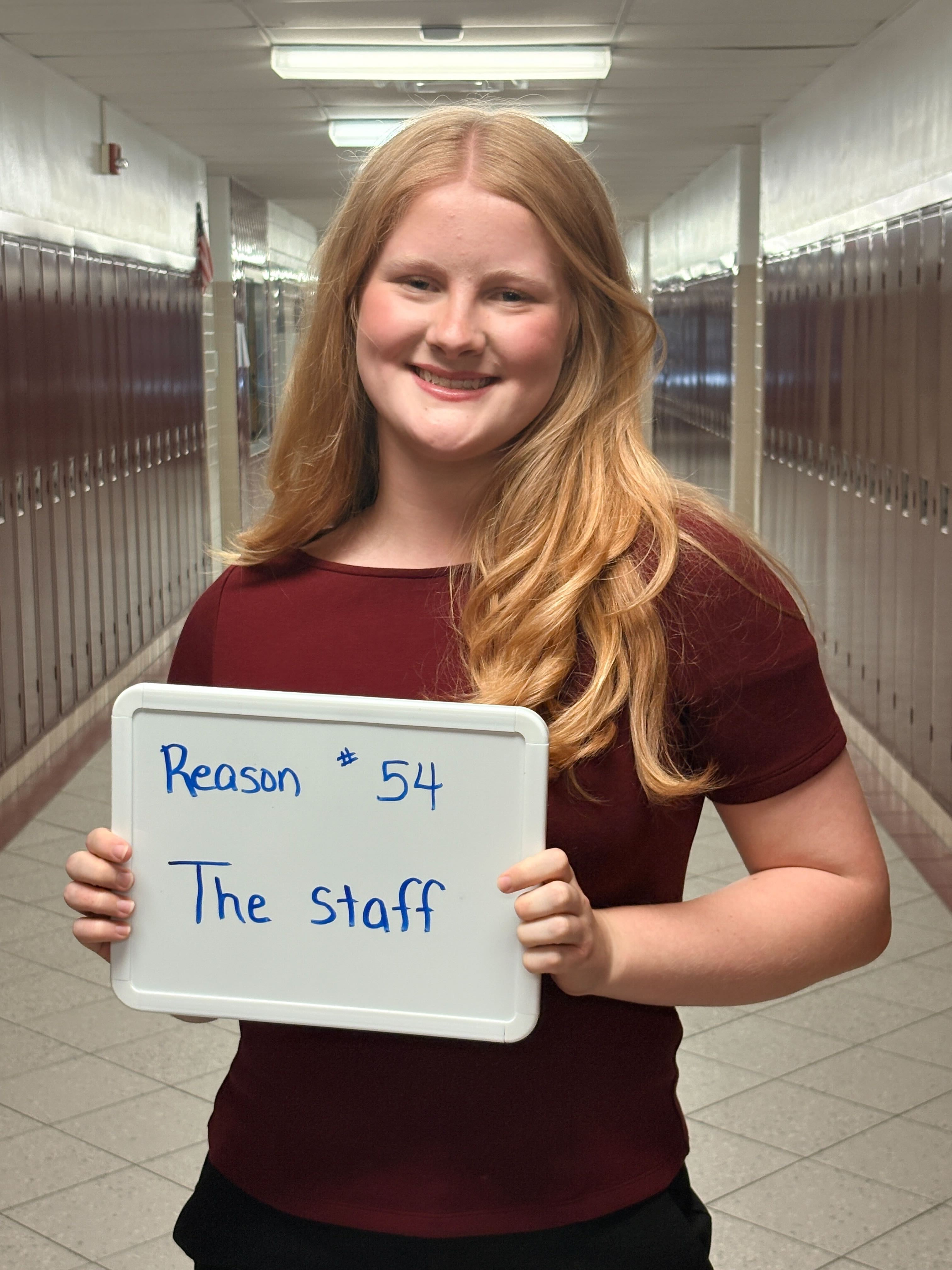 Blonde-haired person in maroon top holds sign with "Reason 54 The staff" on it. They stand in a hallway.