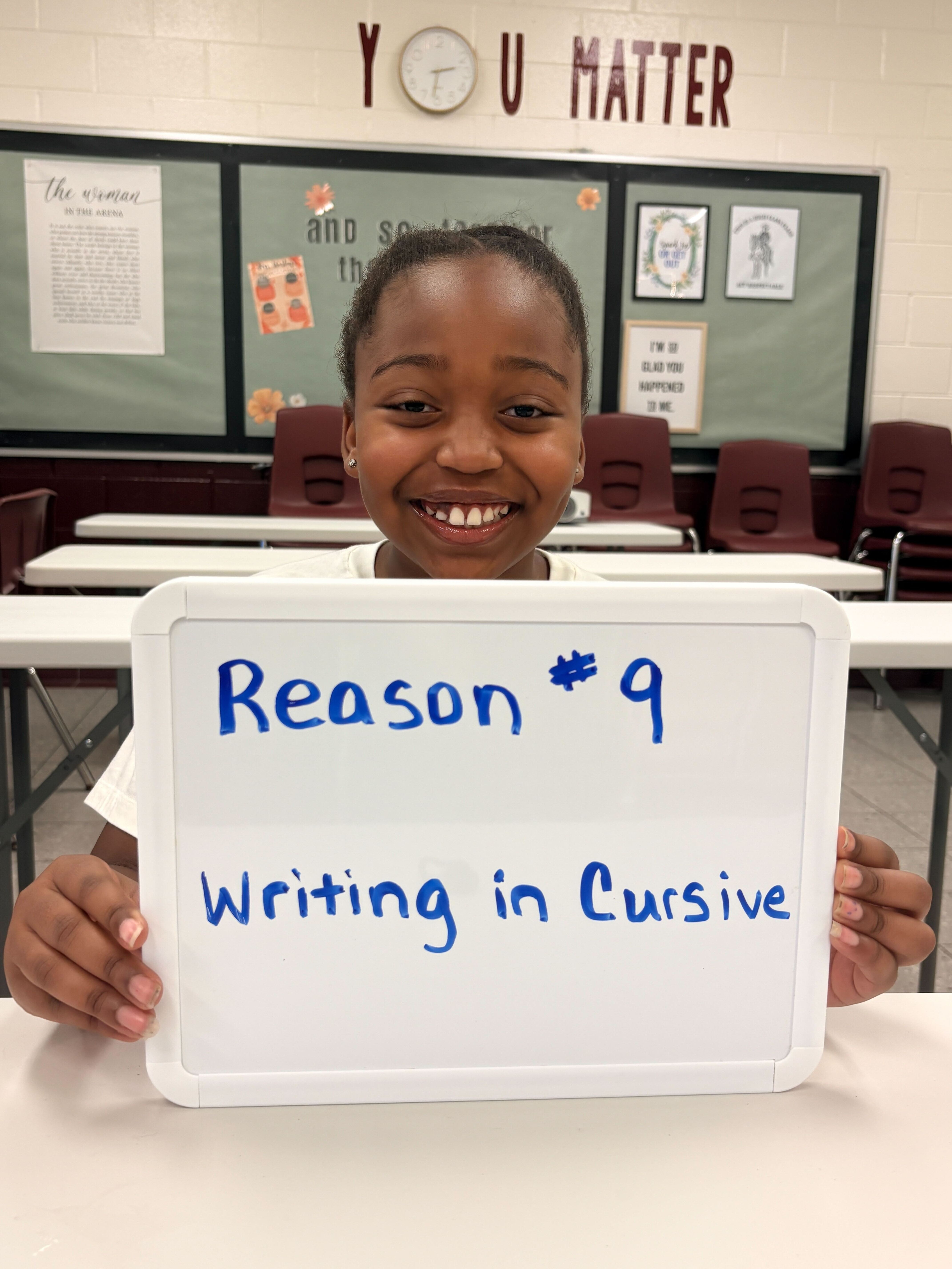 Child in classroom holds sign that reads "Reason #9 Writing in Cursive". Green board with "You Matter" in background.