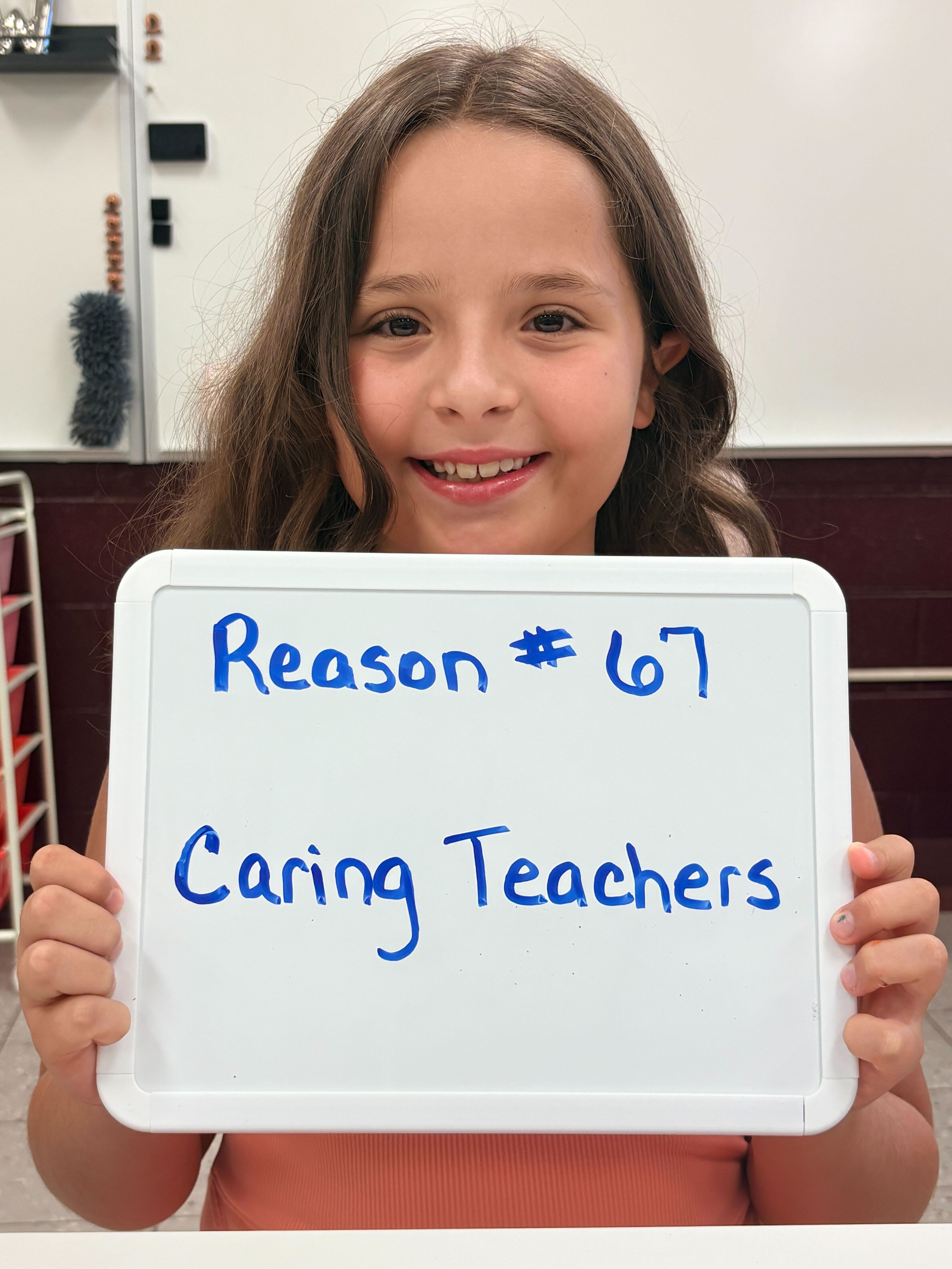 A child holds a white board displaying "Reason #67 Caring Teachers" in a classroom with shelves and a white wall.
