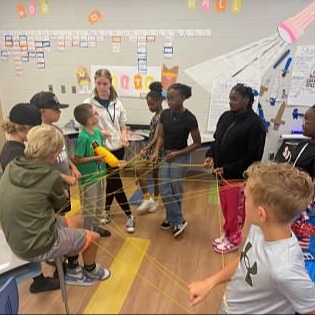 A classroom with children engaged in an activity with yarn or rope, some sitting and others standing.
