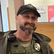 Chris Hayes, School Resource Officer, stands smiling in a law-enforcement style uniform indoors. He wears a black cap, a green jacket with a radio on the shoulder, and a badge pinned to his chest.