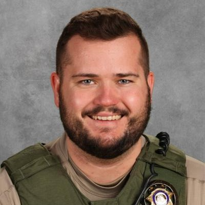Noah Cherry, a School Resource Officer, smiles at the camera while wearing a tan uniform and green tactical vest with a radio microphone attached.
