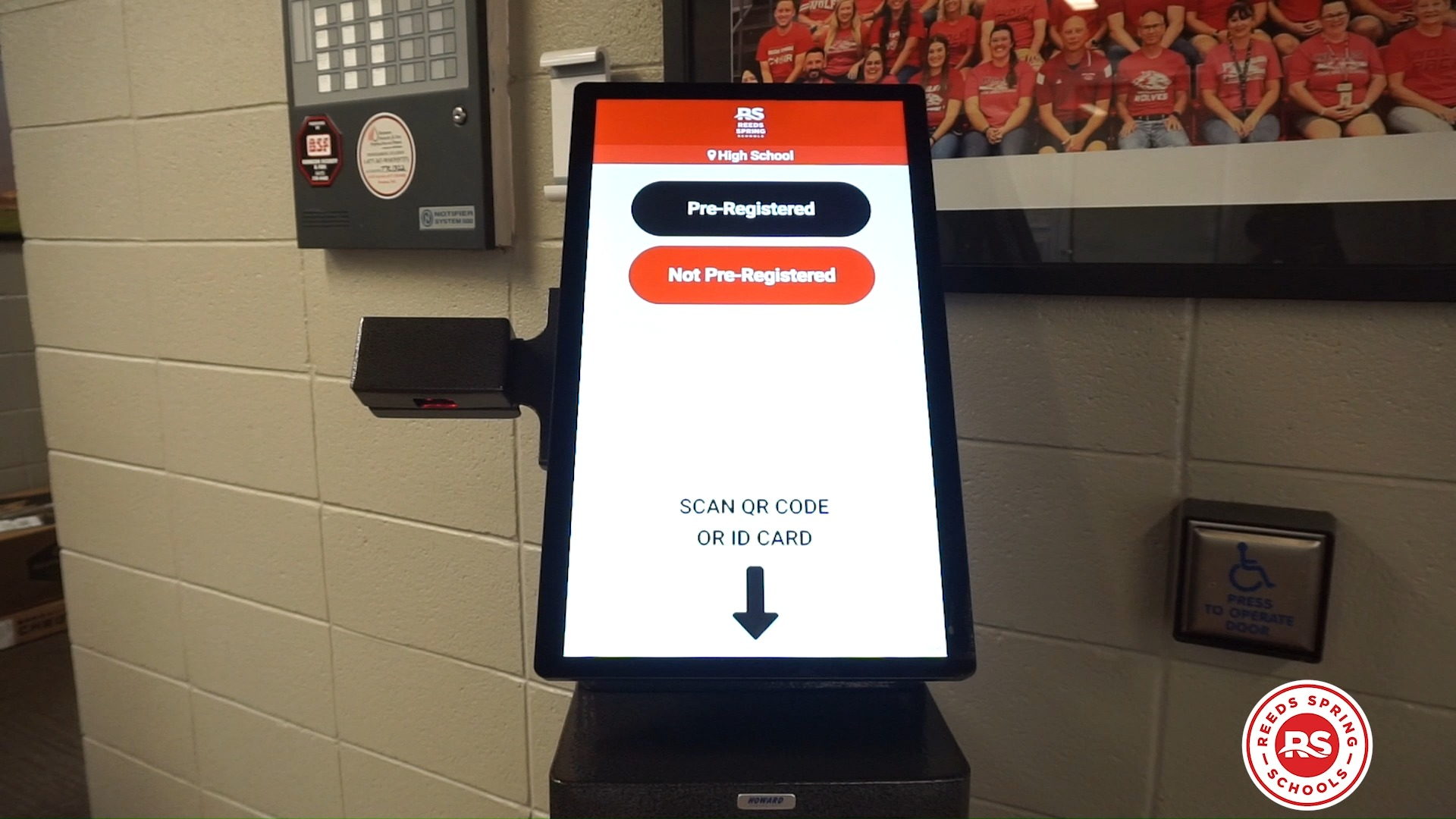 A touchscreen visitor check-in kiosk stands in a school hallway. The screen displays options for pre-registered and not pre-registered visitors above instructions to scan a QR code or ID card.