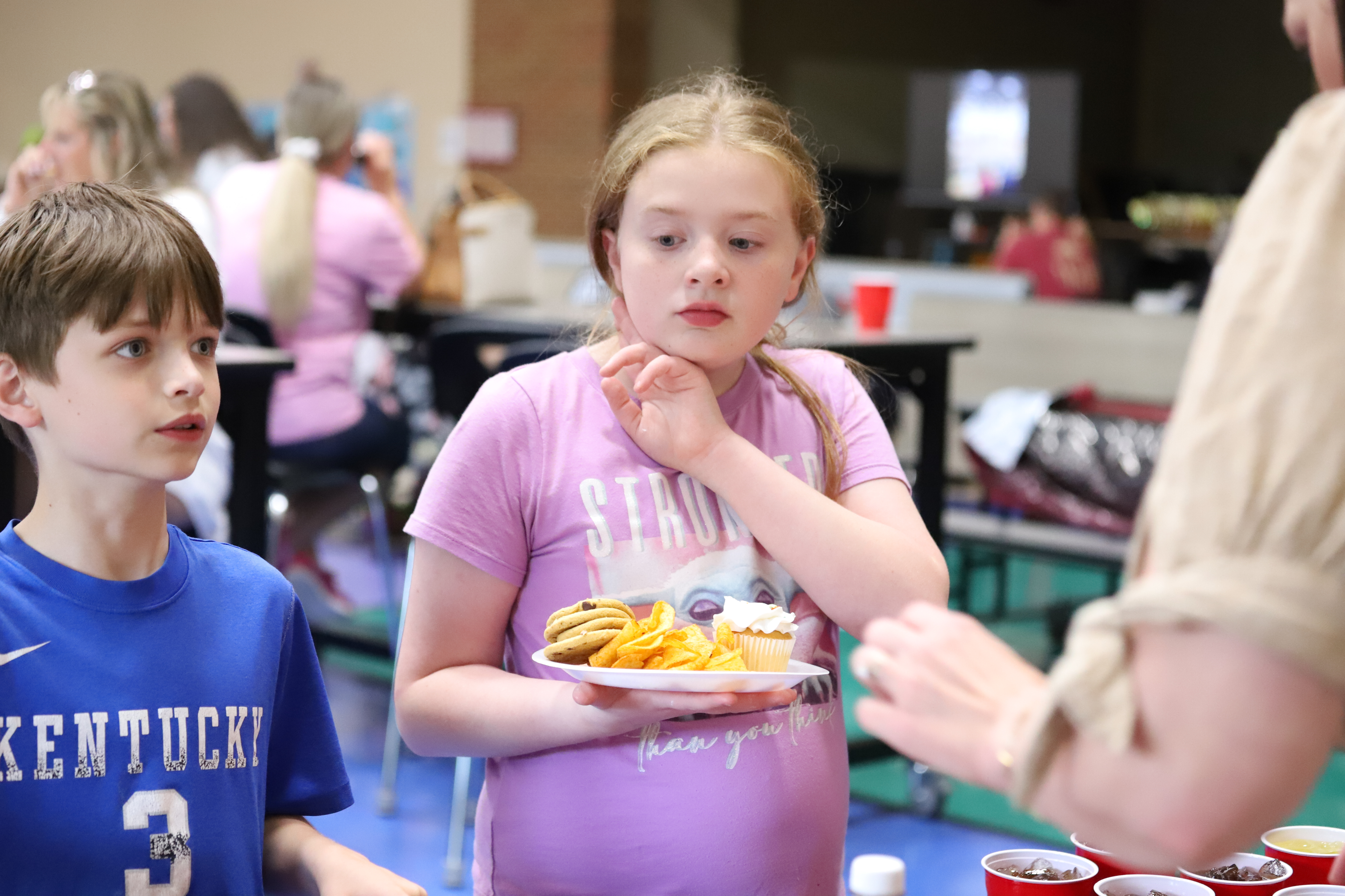 A young girl in a lavender shirt holds a plate of snacks, including cookies, chips, and a cupcake, while standing beside a boy in a blue "Kentucky" t-shirt. They are indoors at what appears to be a casual gathering or event, with people seated at tables in the background. The girl looks slightly to the side with a thoughtful expression as an adult off-camera interacts with them.