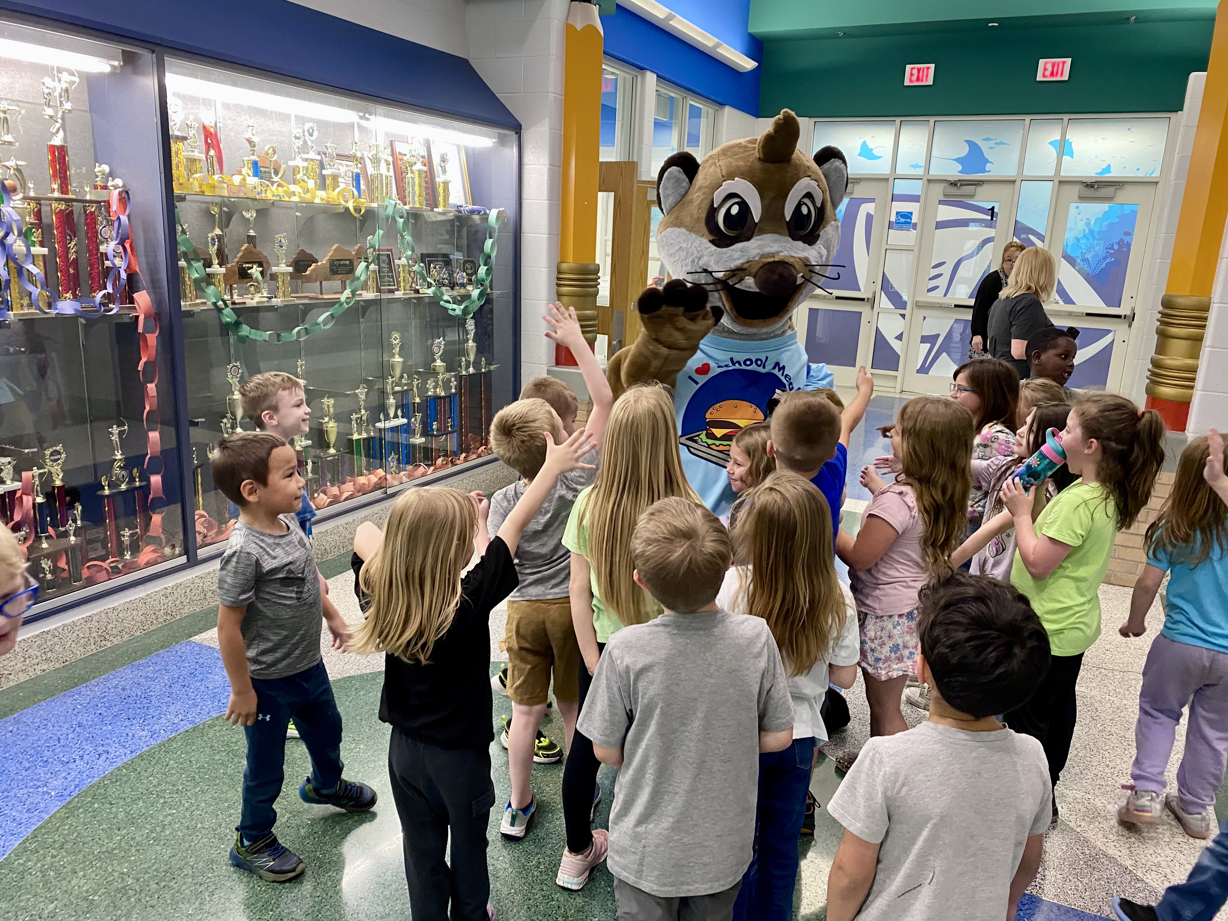 A group of young elementary students gathers excitedly around Mealton the Meerkat mascot in a school hallway. The mascot, wearing a shirt that reads “I ❤️ School Meals,” waves at the camera while children reach out to high-five him. A large trophy case is visible along the wall, adding to the celebratory school environment.
