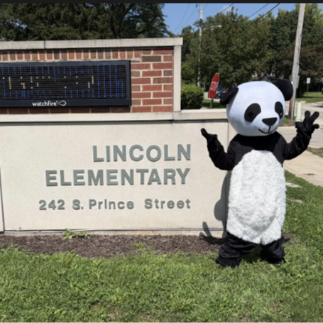 A person in a panda costume stands on grass in front of a brick building with "LINCOLN ELEMENTARY" sign.